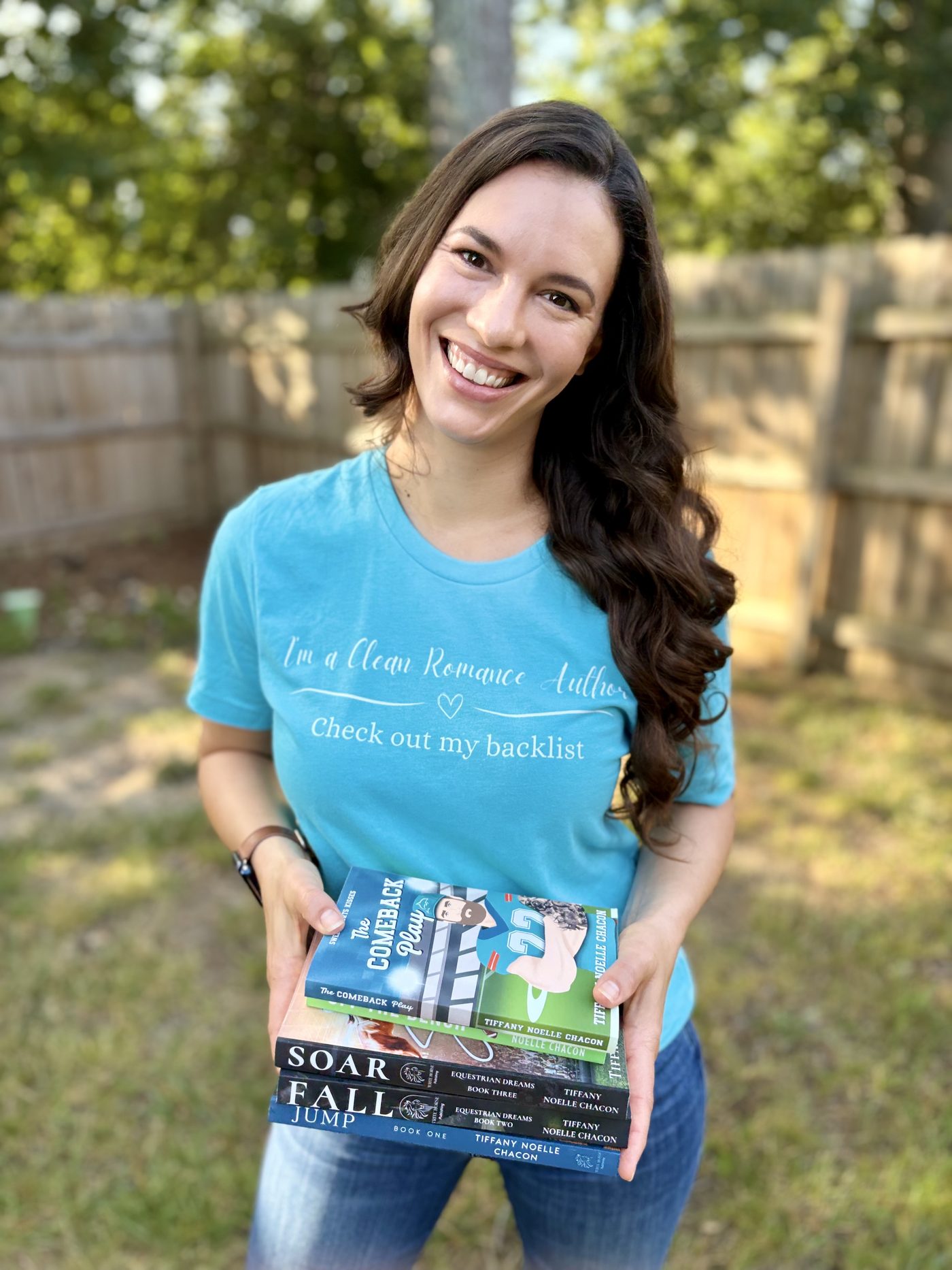 Tiffany Noelle Chacon smiling outdoors, holding a stack of her clean romance novels, wearing a turquoise 'I'm a Clean Romance Author' T-shirt